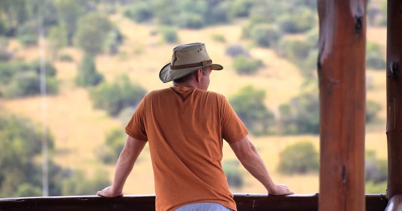 A man in a sun hat resting on a wooden railing, gazing calmly out over a warm sunlit valley