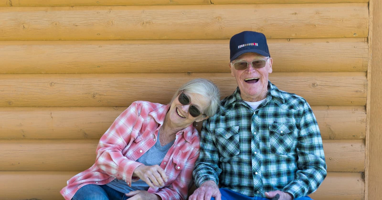 An older couple sitting close together against a log wall, laughing warmly in the sunshine
