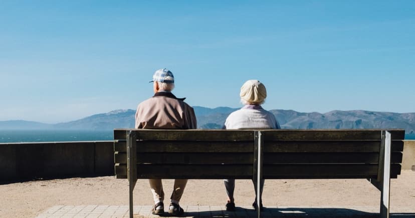 A retired couple sitting together on a bench, looking out at the sea and distant mountains